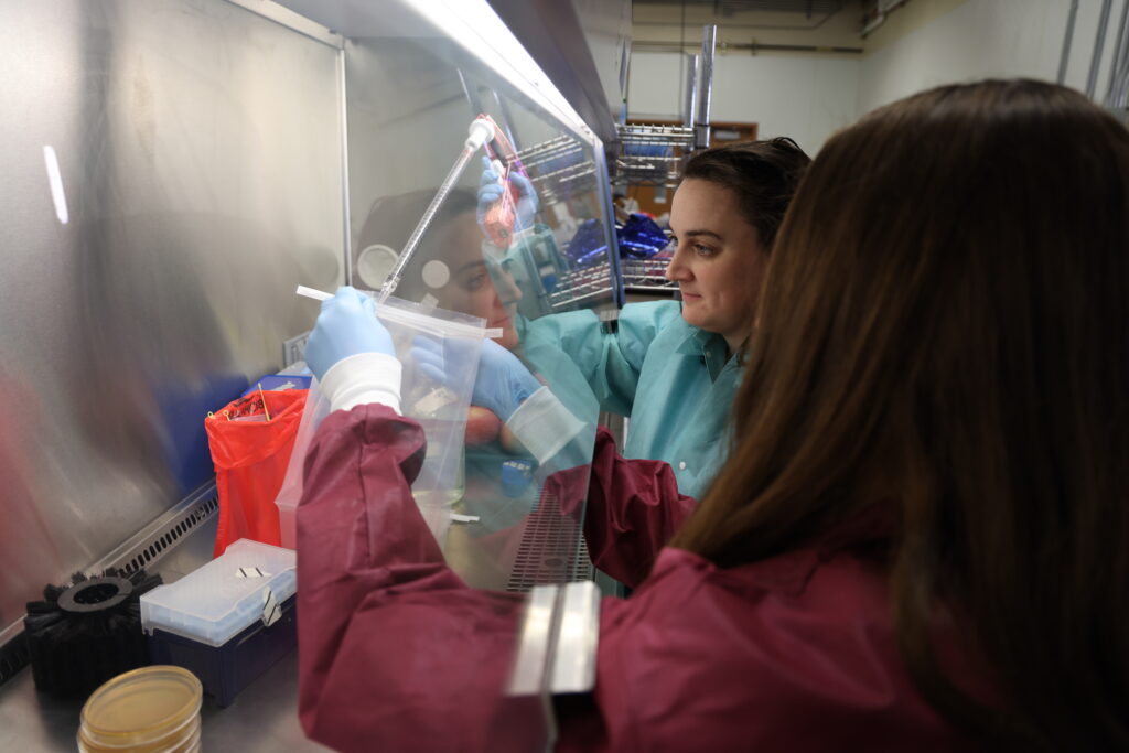 Murphy, Center, and Andreson, with back to camera, in protective suits and gloves, handling materials and cylinders under a glass hood.
