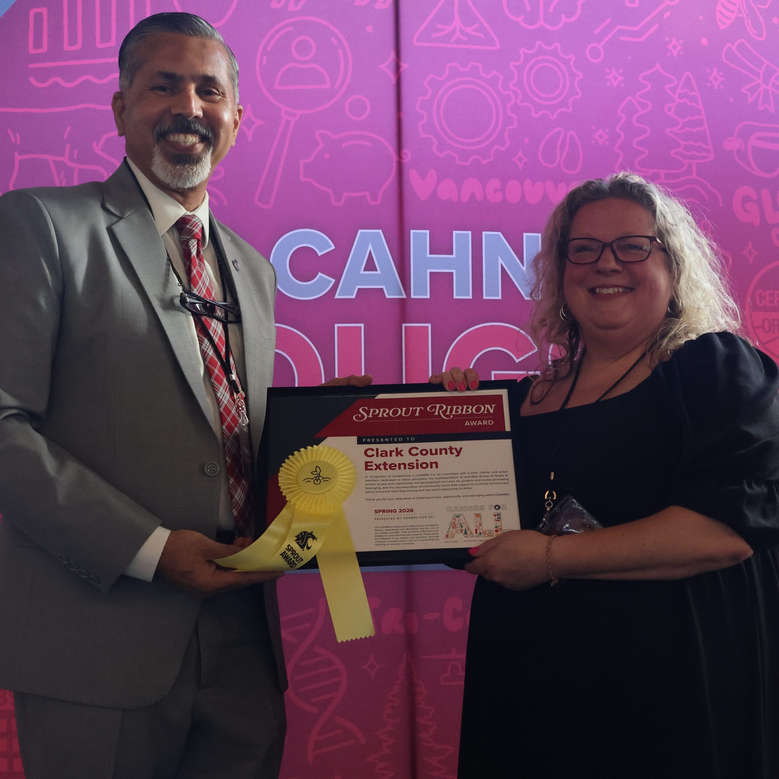 Ceremony participant Missy McElprang, right, holds an award certificate on stage in front of WSU-themed backdrop. CAHNRS Dean Raj Khosla stands at left, helping hold the certificate.