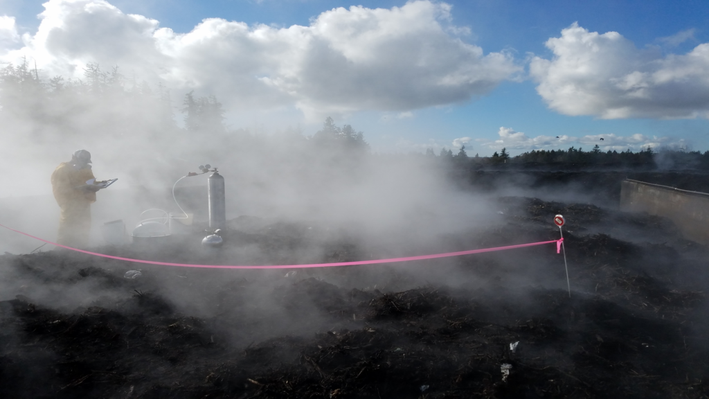 A person in yellow protective gear and a gas mask stands next to scientific equipment on a hill that is emitting lots of steam. There is a red tape barrier around the mound.