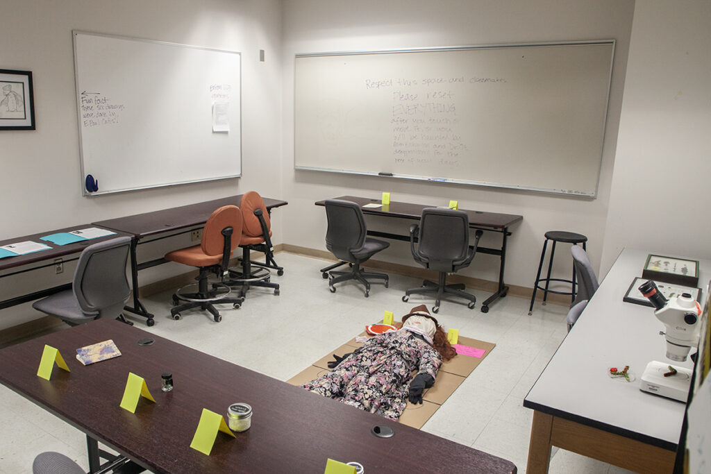 A windowless large room with a stuffed body in a dress laying on the floor. Tables are spread out in a square around the body with different crime scene clues on each.