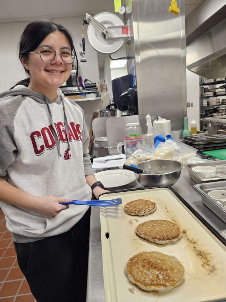 A person wearing a WSU sweatshirt holds a spatula while standing over a griddle that has 3 pancakes cooking on it. The background looks like an industrial kitchen.