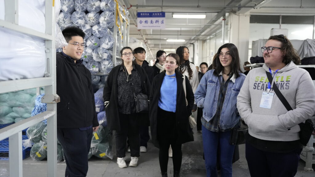 9 people stand in a factory, with dozens of rolls of fabric behind them.