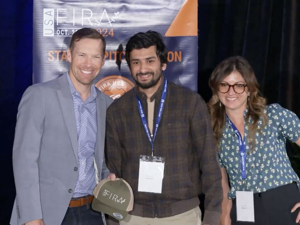 Three people stand together. One holds a hat with the letters “USA FIRA" on it. Behind them is a banner that says “USA FIRA” and other words that are obscured.