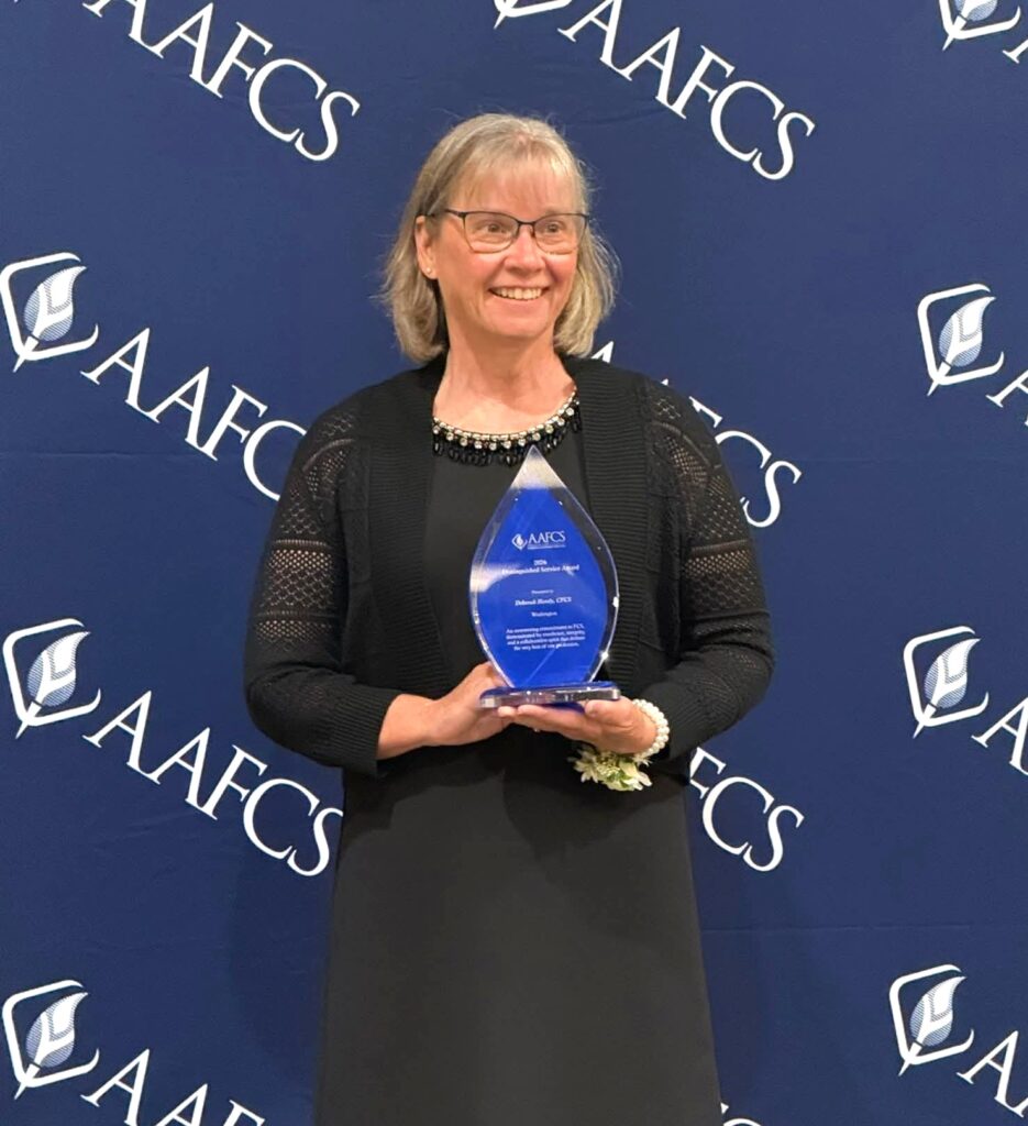 Debbie Handy holds an award while smiling and standing in front of a background with the acronym AAFCS and the AAFCS logo repeated across it.