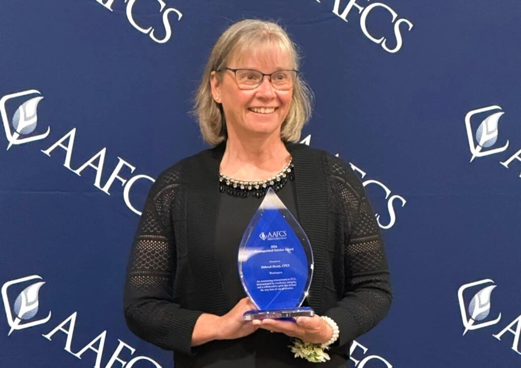 Debbie Handy holds an award while smiling and standing in front of a background with the acronym AAFCS and the AAFCS logo repeated across it.