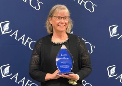Debbie Handy holds an award while smiling and standing in front of a background with the acronym AAFCS and the AAFCS logo repeated across it.