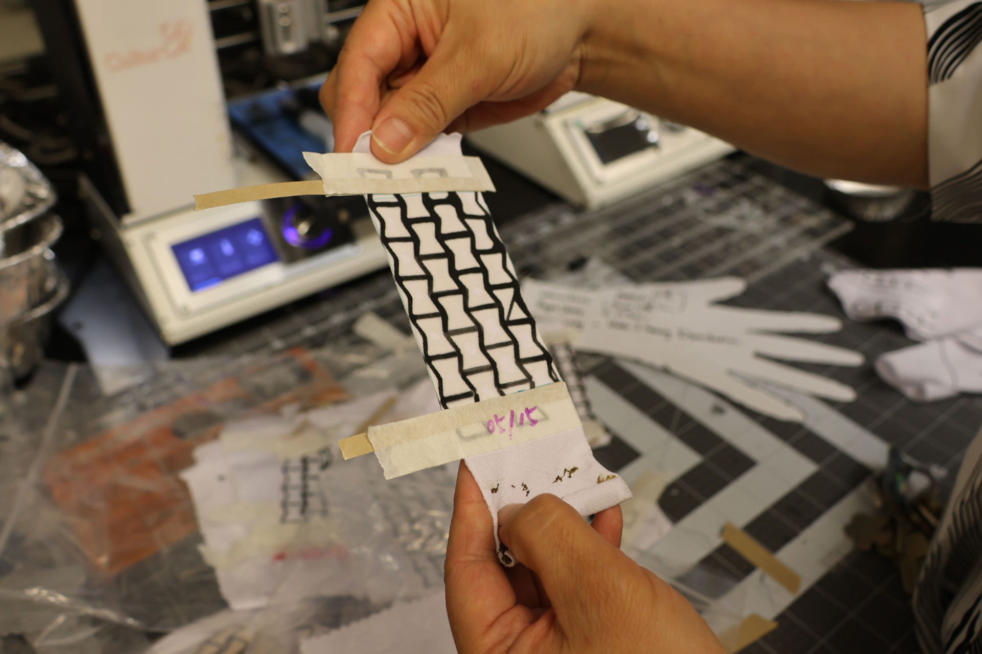 Hands holding a swatch of white fabric with a lattice of black lines printed atop it, in a lab.