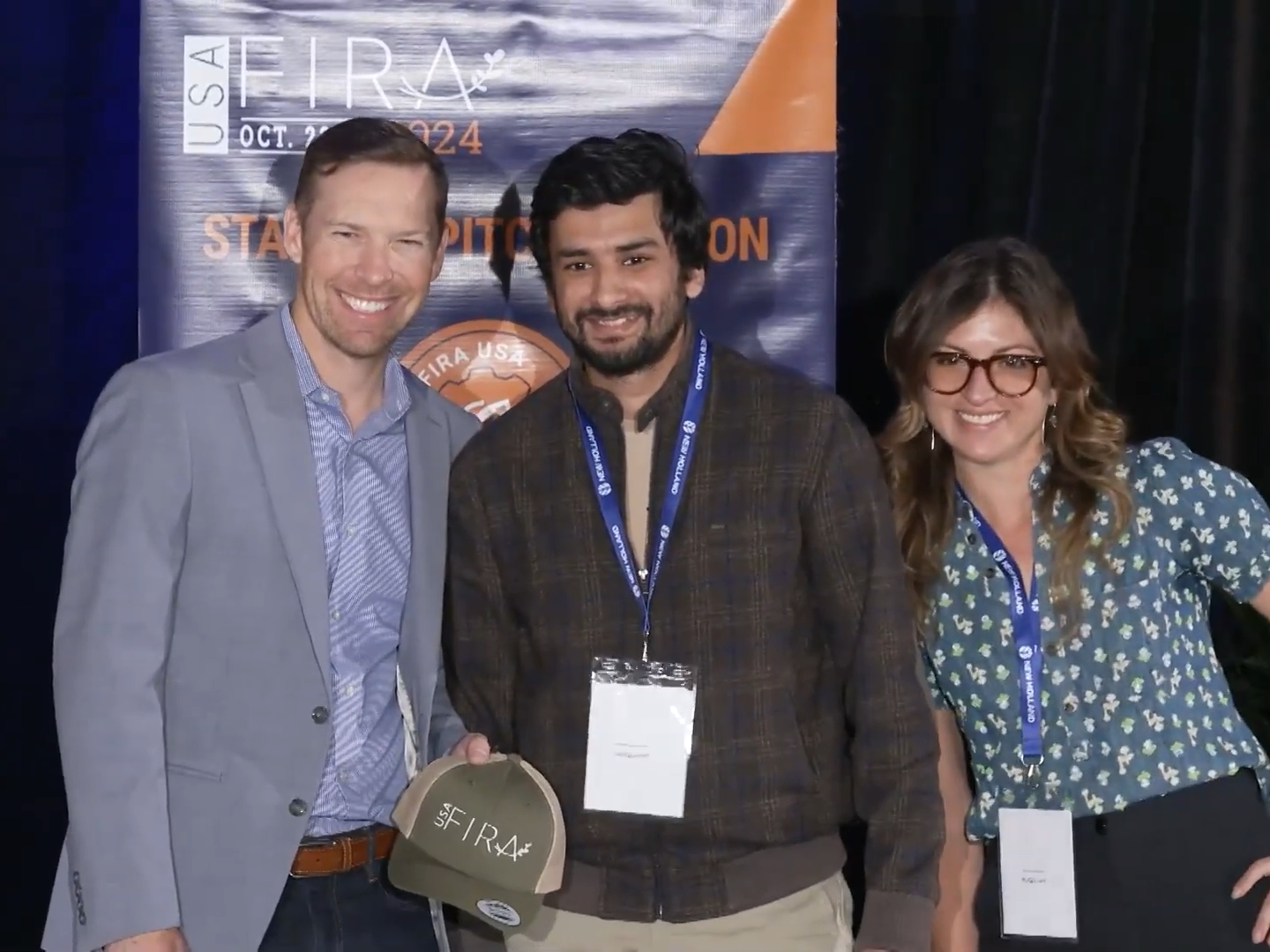 Three people stand together. One holds a hat that says "USA FIRA" on it . Behind them is a banner that says USA FIRA and other words that are obscured.