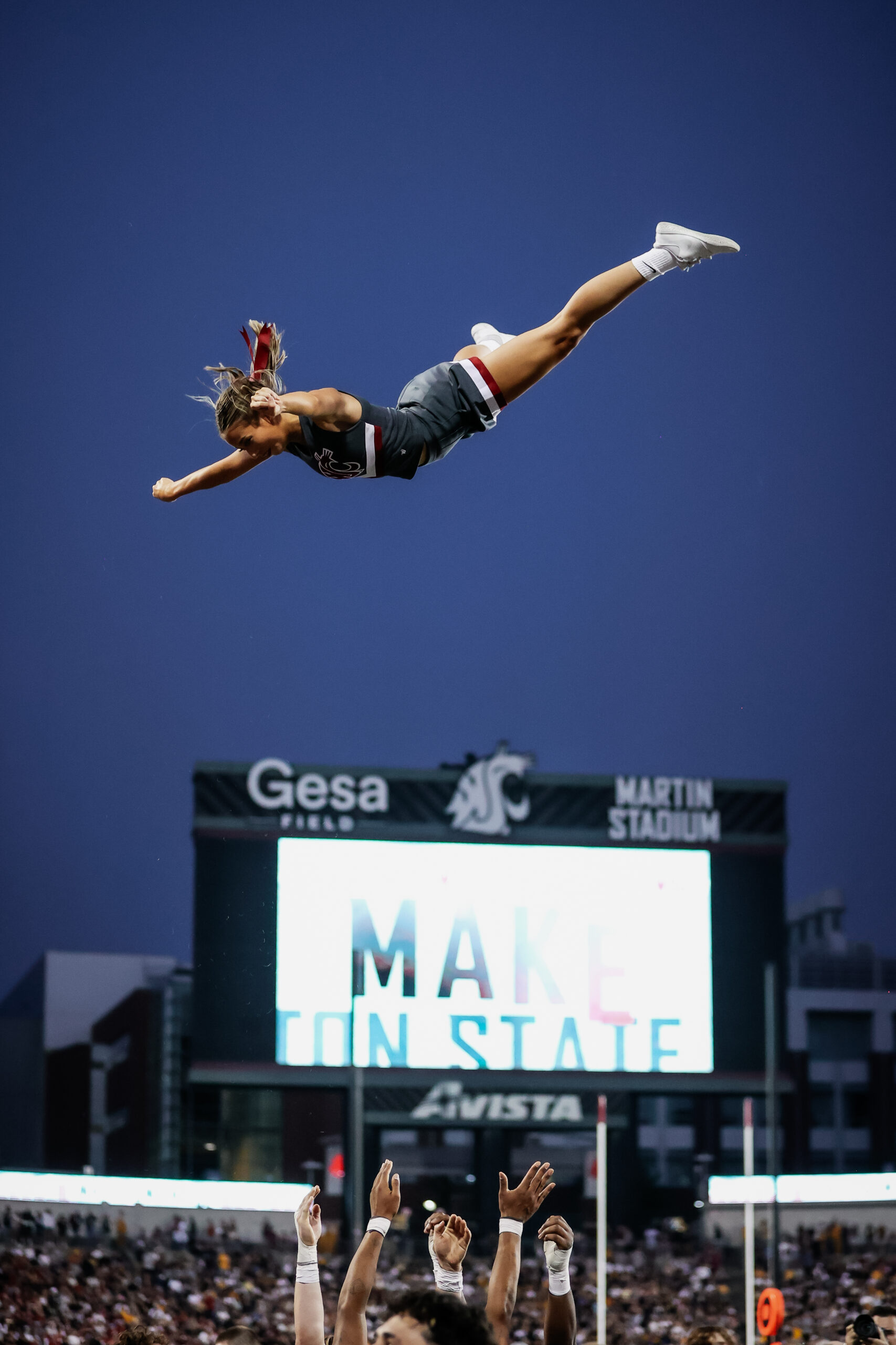 A person is tossed into the air, horizontal to the ground. It appears she’s above a large scoreboard in the background. Several hands below her reach up to catch her.