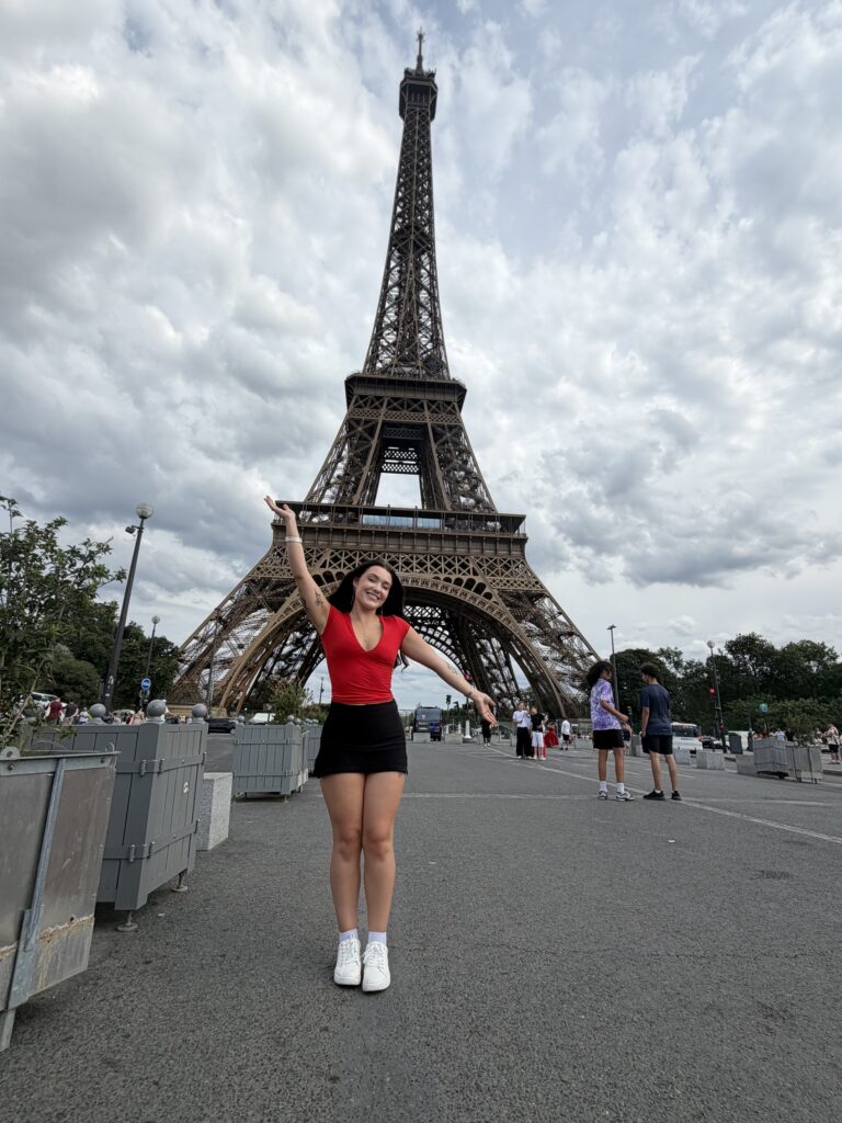 A woman raises one arm to emphasize the Eiffel Tower behind her.