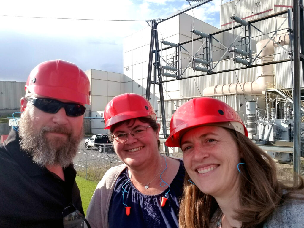 3 people wearing orange hard hats stand outside an industrial-looking facility.