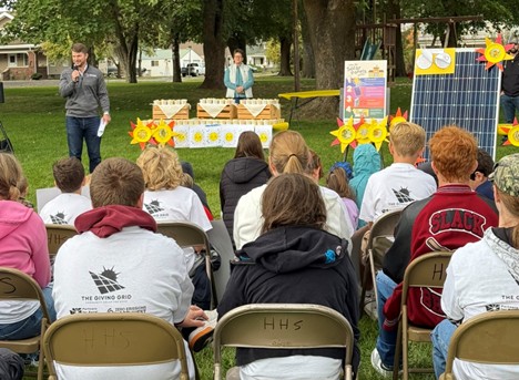 A group of students face a speaker in a park-like setting. A solar panel and posters are propped up to the side of the speaker.