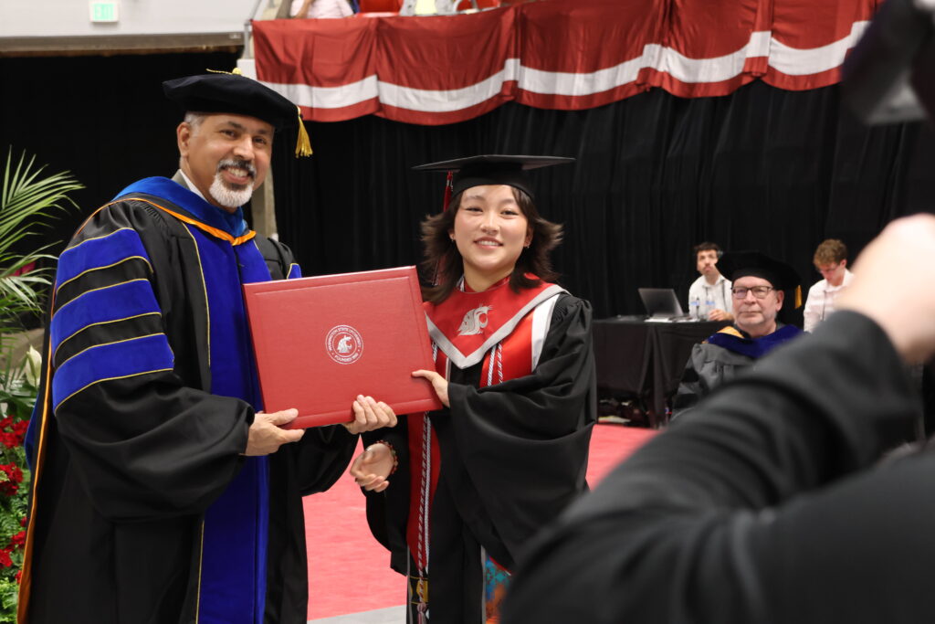Khosla and Grosz, in commencement robes, smile while holding diploma in front of a photographer