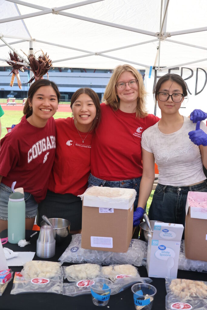 Four students smile under an awning with ice cream boxes, spoons, cups, and a water bottle in front of them on a table.