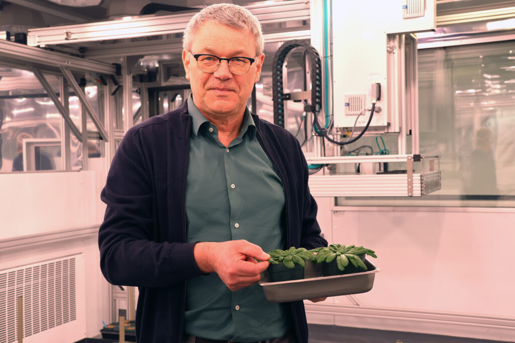 Kirchhoff, standing, holding small tray of plants in front of scientific instrument.