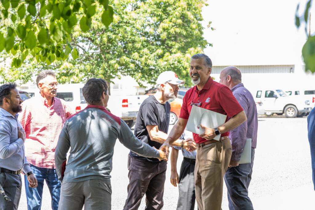 Dean Kholsa, smiling, shakes hands with a colleague as others gather, shake hands, and smile, outdoors.