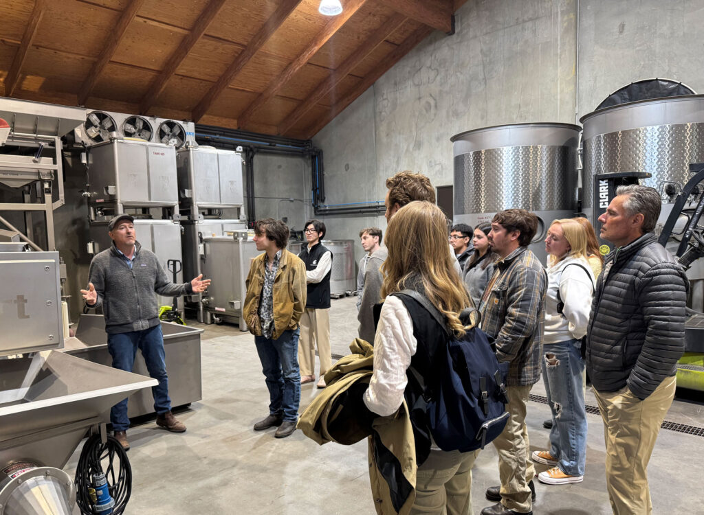 A group of about a dozen people stands next to metal wine tanks and winemaking equipment in a concrete building with wooden paneled ceilings.