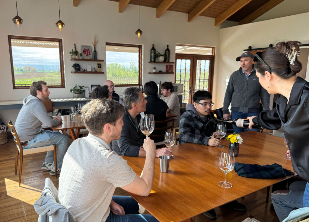 Five people sit at one table, and three sit at another. Both tables have wine glasses on them. A person stands while pouring wine into a glass at one of the tables. Another person stands near the table.