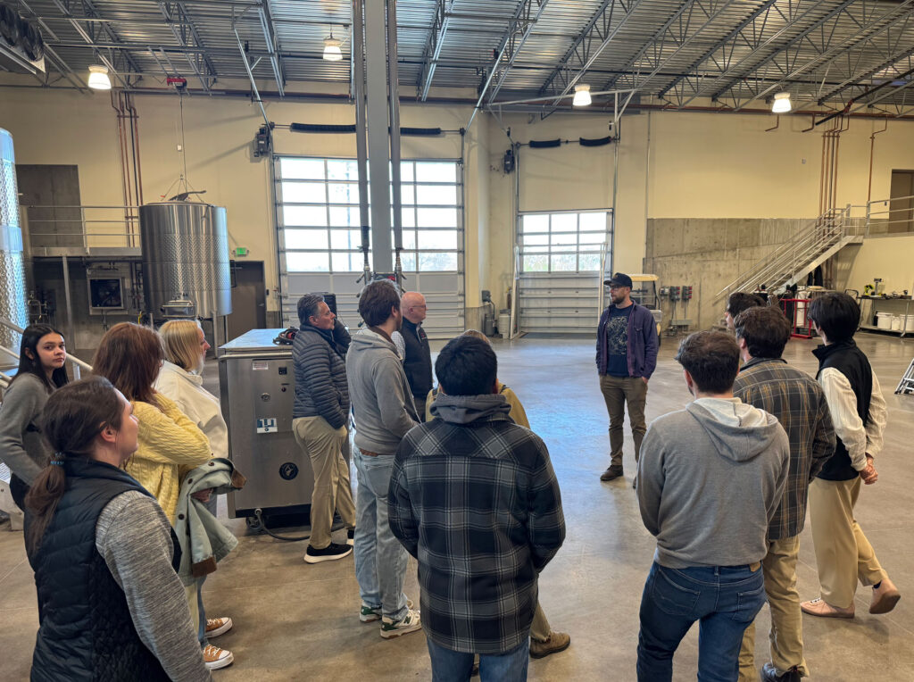 A group of about 14 people stands in a warehouse with metal wine tanks and other equipment.
