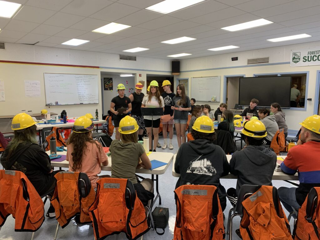 A group of teens, some in hard hats and personal protective equipment, attend an in-classroom presentation.