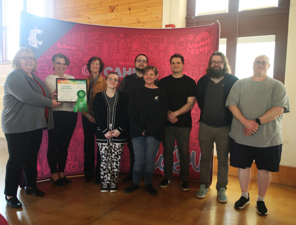 A group of nine people stands in front of a WSU CAHNRS-themed background. The two people on the left hold an award with a ribbon attached to it.