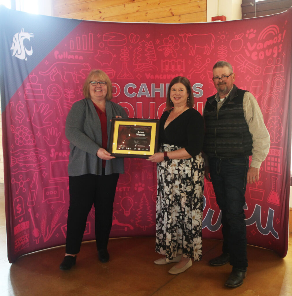 Three people stand together in front of a WSU CAHNRS-themed background. The two on the left hold an award.