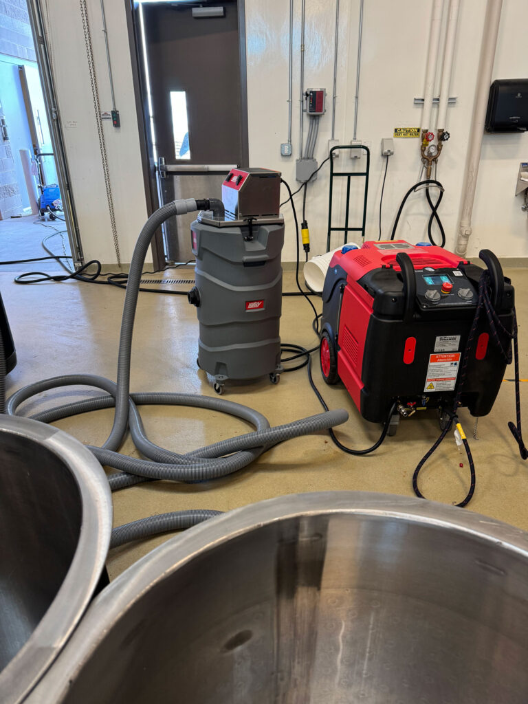 A large vacuum sits on the floor near steel wine tanks and a piece of equipment with a hose coming out of it.