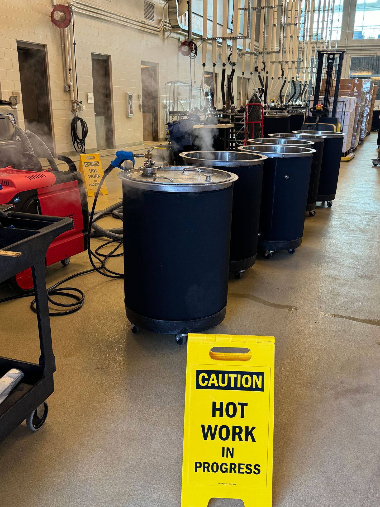Several wine fermentation tanks sit in a row. One has steam coming out of it. Next to the tanks is a large vacuum-like piece of equipment, and a rolling cart. IN ther background is other winemaking equipment. Signs in front and back of the tanks reads "Caution hot work in progress."