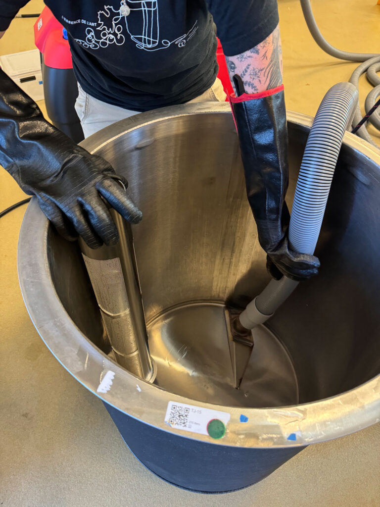 A person's gloved hands hold a vacuum-like hose inside of a steel wine tank.