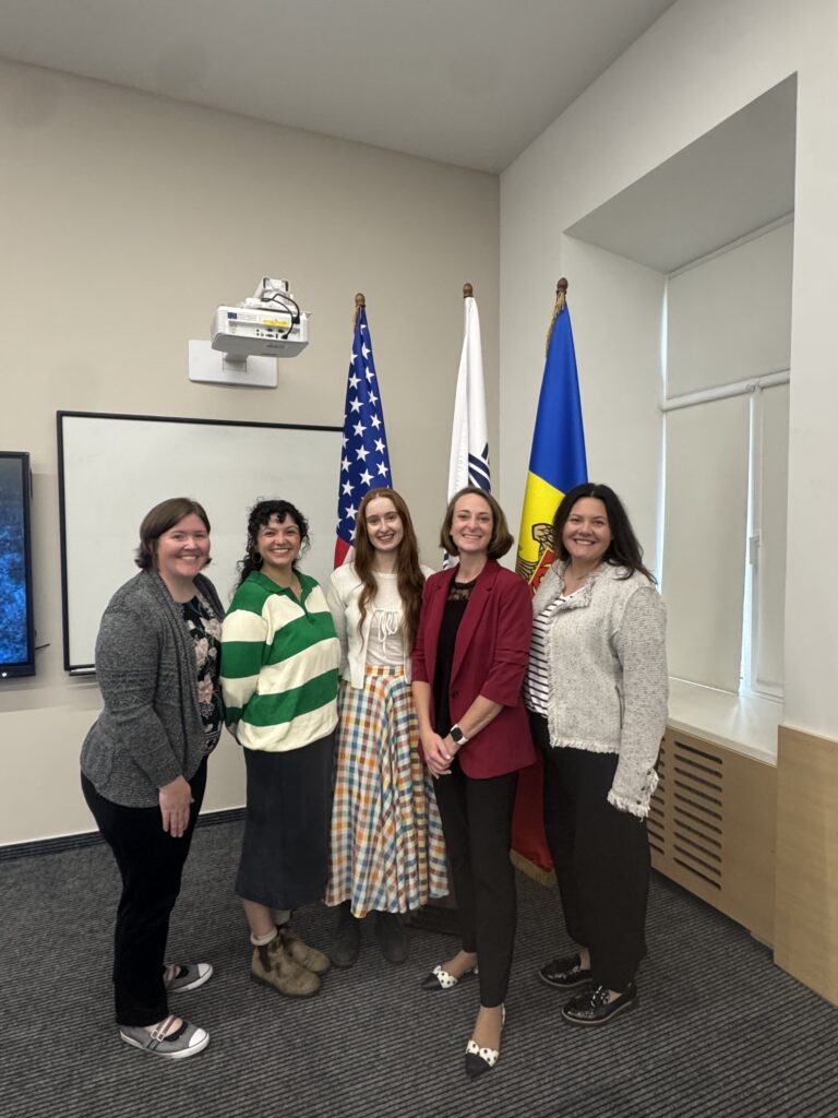 A group of five people stands in front of three flags.
