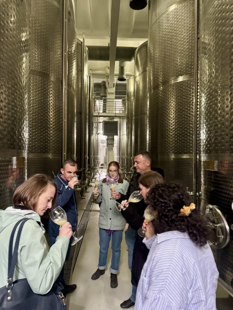 A group of six people smells and tastes glasses of wine while standing next to large metal wine tanks.