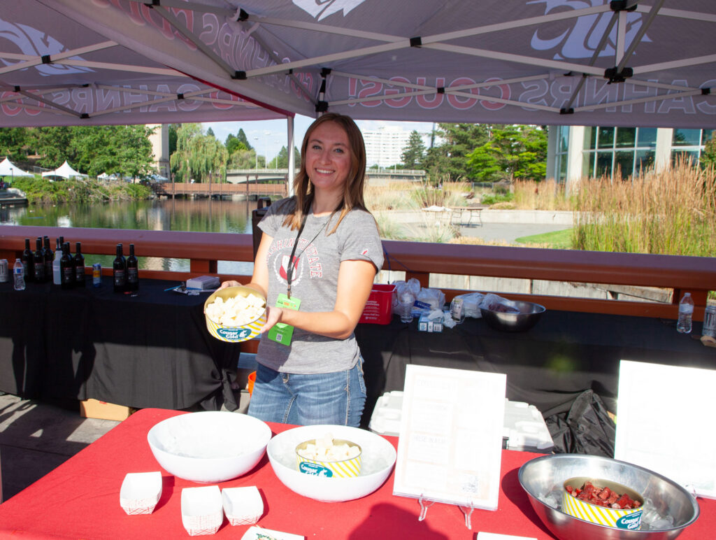 A person holds a can of Cougar Gold cheese samples while standing under a CAHNRS themed tent. On the table in front of her are another can of cheese samples in a bowl of ice and a can of beef stick samples in a bowl of ice.