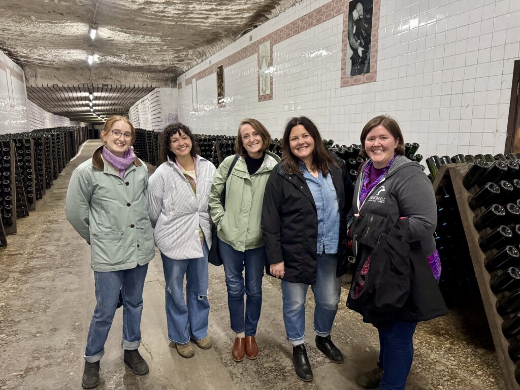 A group of five people stands in a room with white tiles on the wall. Nearby, there are many wooden racks filled with wine bottles.