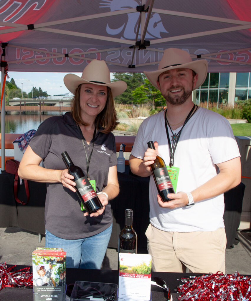Two people hold bottles of red wine while standing under a CAHNRS themed tent. On the table is another bottle of wine, pom poms, and brochures about WSU's wine science program.