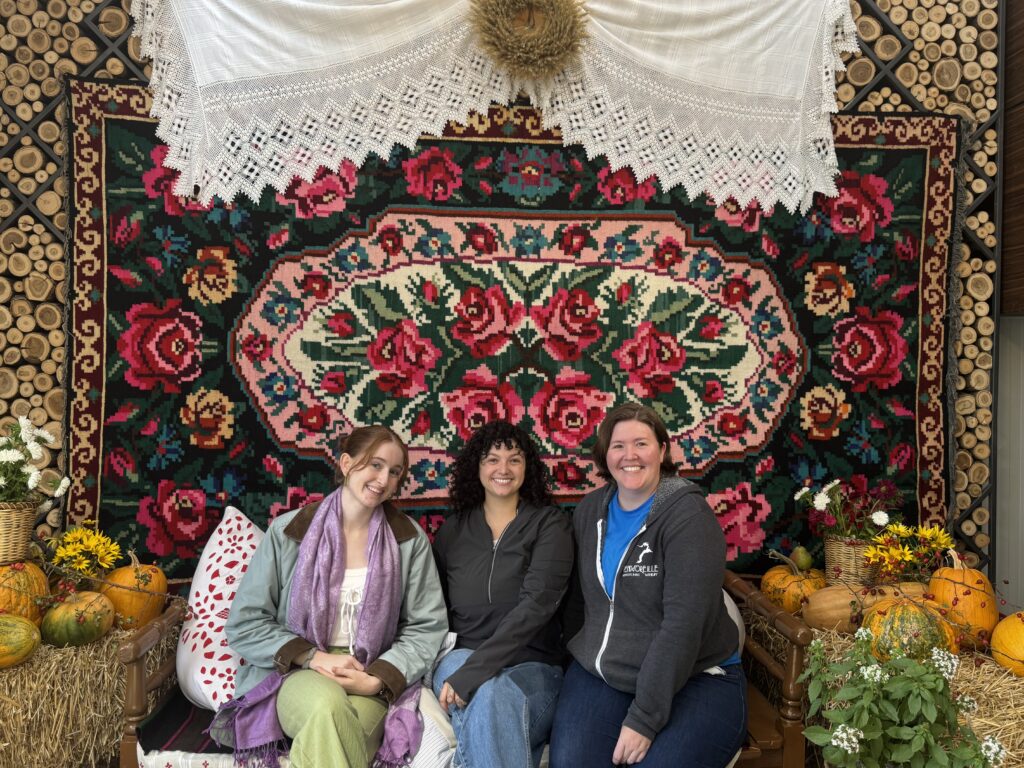 Three people sit in front of a colorful flowered rug that is hanging over many rows of stacked wooden logs that have just the ends visible. Above the rug is a white fabric hanging with a decorative lace edge. In the middle of the white fabric hanging is a wreath. Next to the group, on either side, are pumpkins, squash, flowers, and other plants.