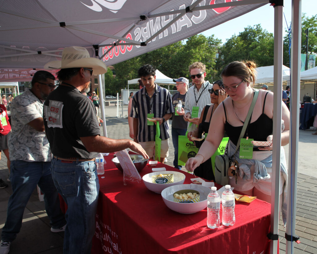 Four people stand in front of a table, while two people stand behind it. On the table are cans of cheese samples in bowls of ice and a can of beef sticks in a bowl of ice.