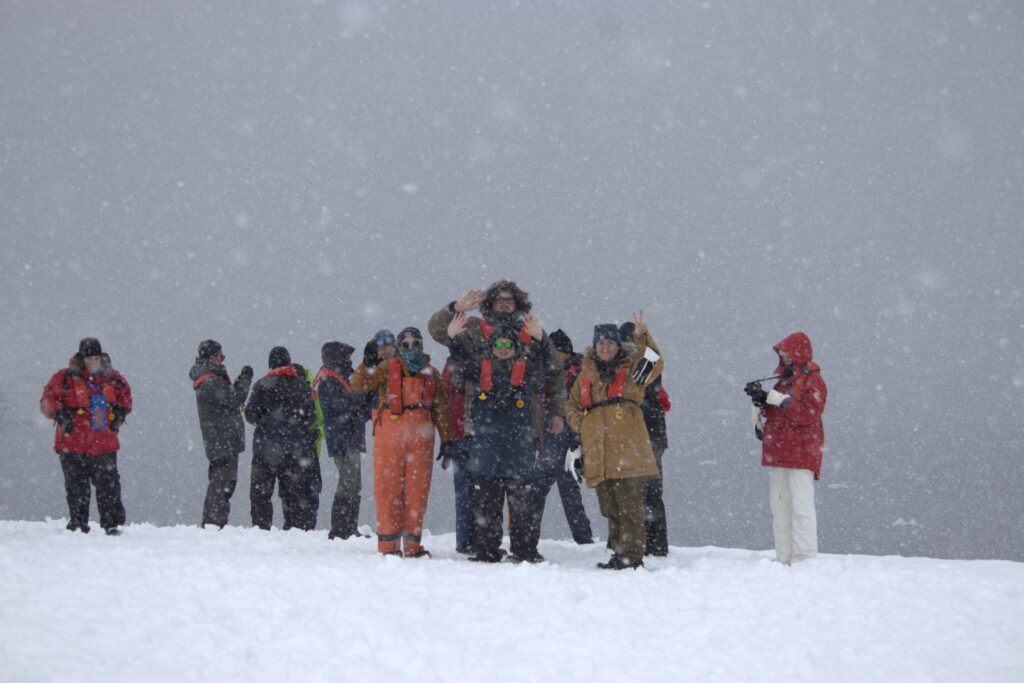 Around a dozen people stand on snow-covered land as snow comes down. 4 of them are waving at the camera.