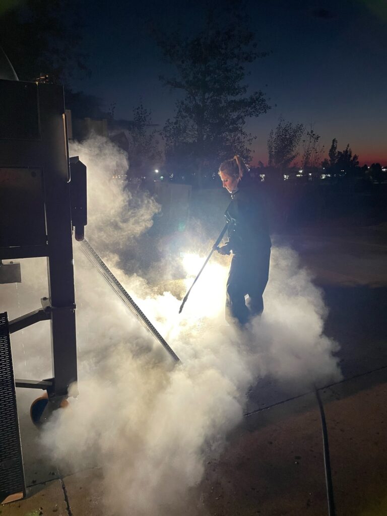 At night, a person stands outside as steam or smoke rises from the ground. They are next to equipment and they are holding a long pole-like tool.