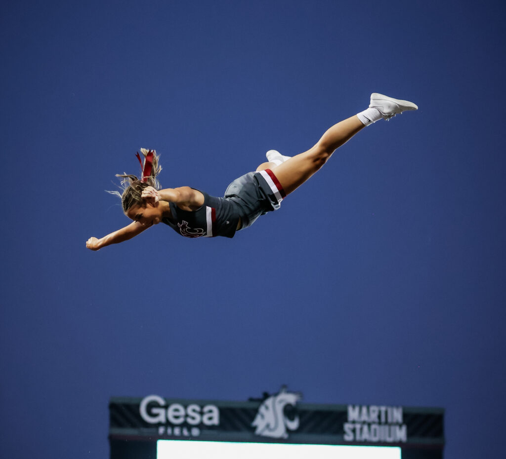 A person is tossed into the air, horizontal to the ground. It appears she’s above a large scoreboard in the background. Several hands below her reach up to catch her.