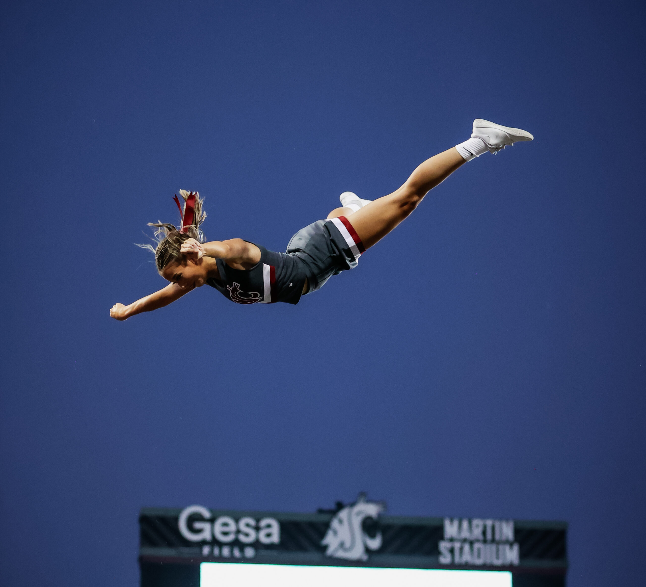 A person is tossed into the air, horizontal to the ground. It appears she’s above a large scoreboard in the background. Several hands below her reach up to catch her.