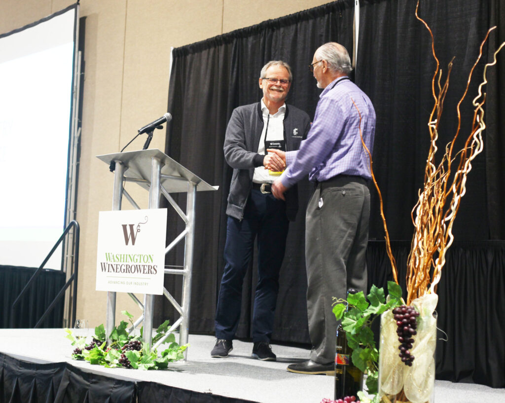 Two people shake hands while one receives an award. They are standing on a stage next to a podium with a sign that reads "Washington Winegrowers. Advancing our industry." There are fake plant decorations on the stage.