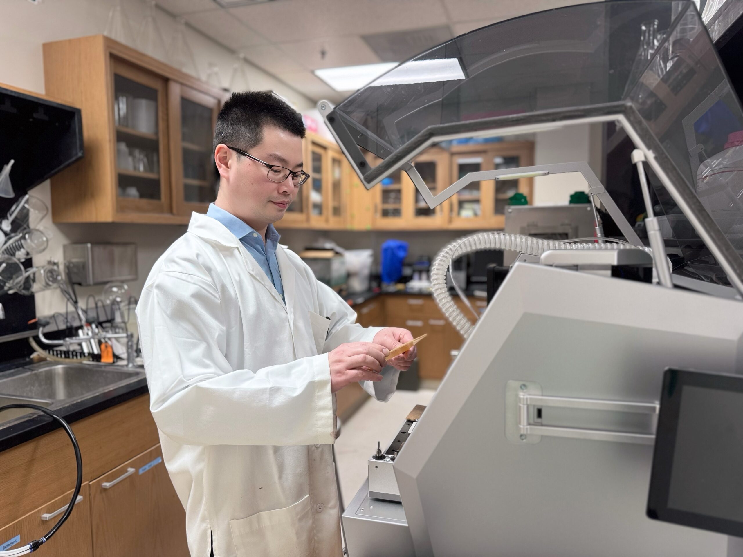 A person in a white lab coat holds a small item while standing in front of a large piece of scientific machinery.
