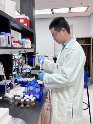 A person in a white lab coat uses scientific equipment other to put material from one item into a test tube in a laboratory.