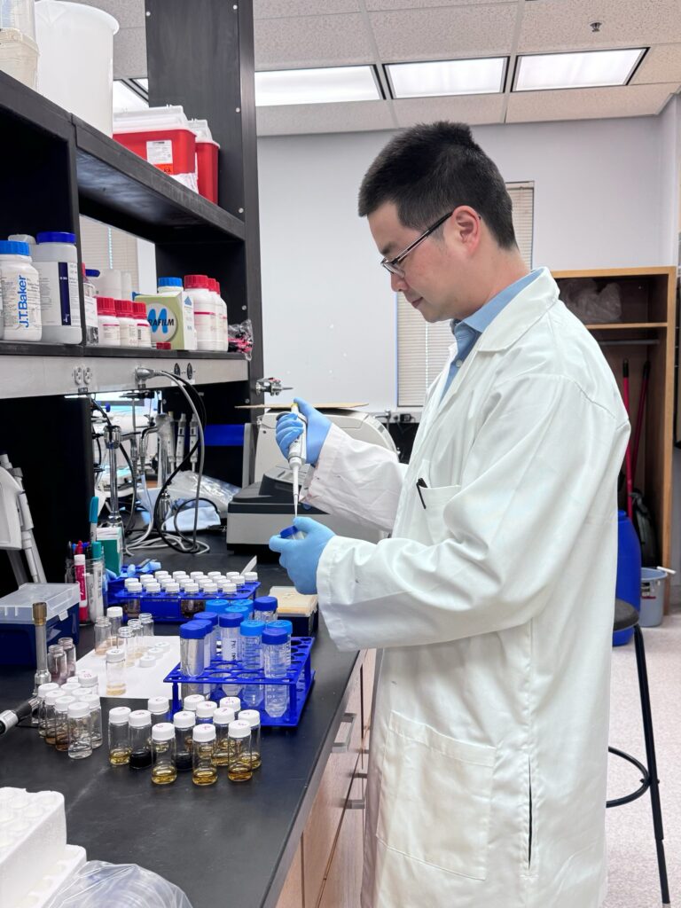 A person in a white lab coat uses scientific equipment other to put material from one item into a test tube in a laboratory.
