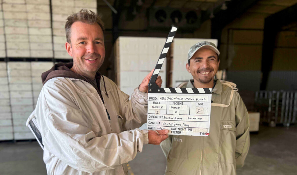 Miller, left, holds open clapperboard, as Ramirez smiles at right, in front of pallet stacks of boxes and containers in a large warehouse room.