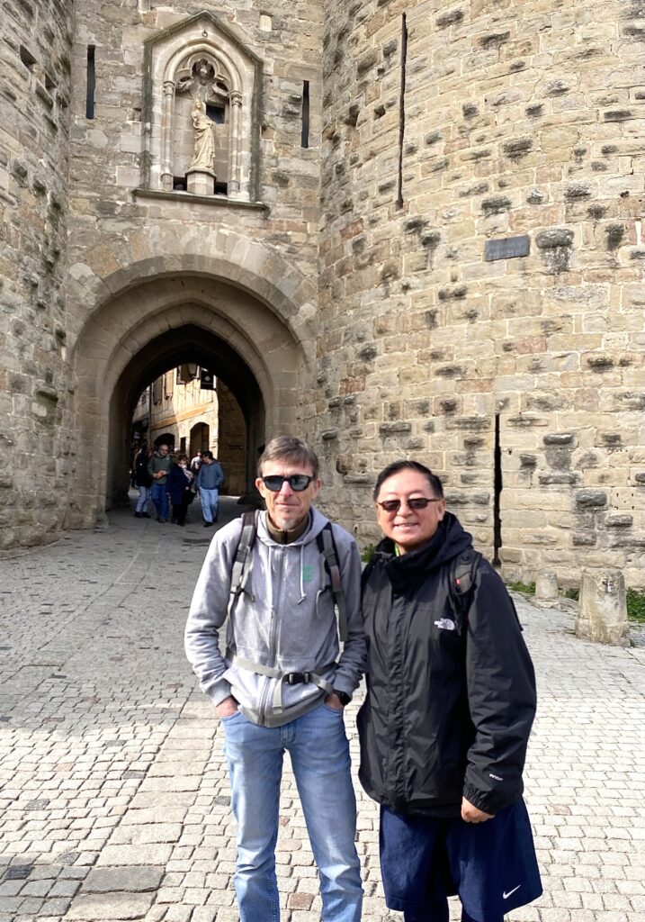 Two men, standing outside a stone gateway on a stone-paved street.
