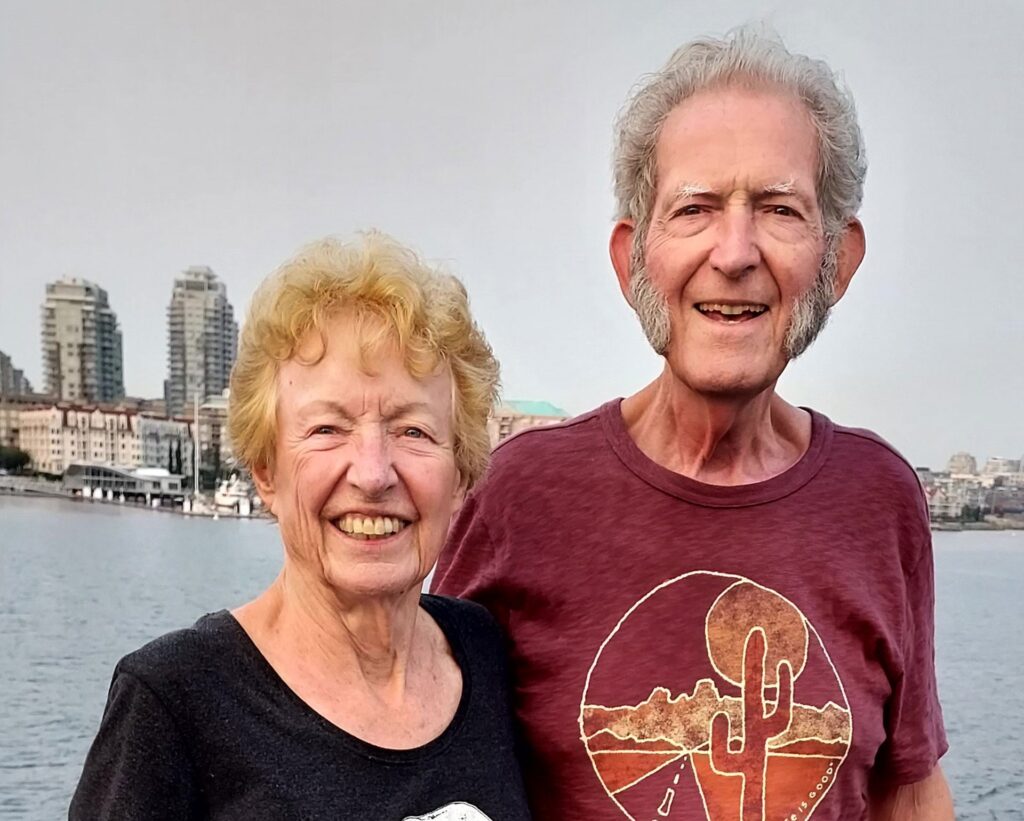 Sue Fairbanks and Jim Huff stand next to the water. A city skyline is in the background.