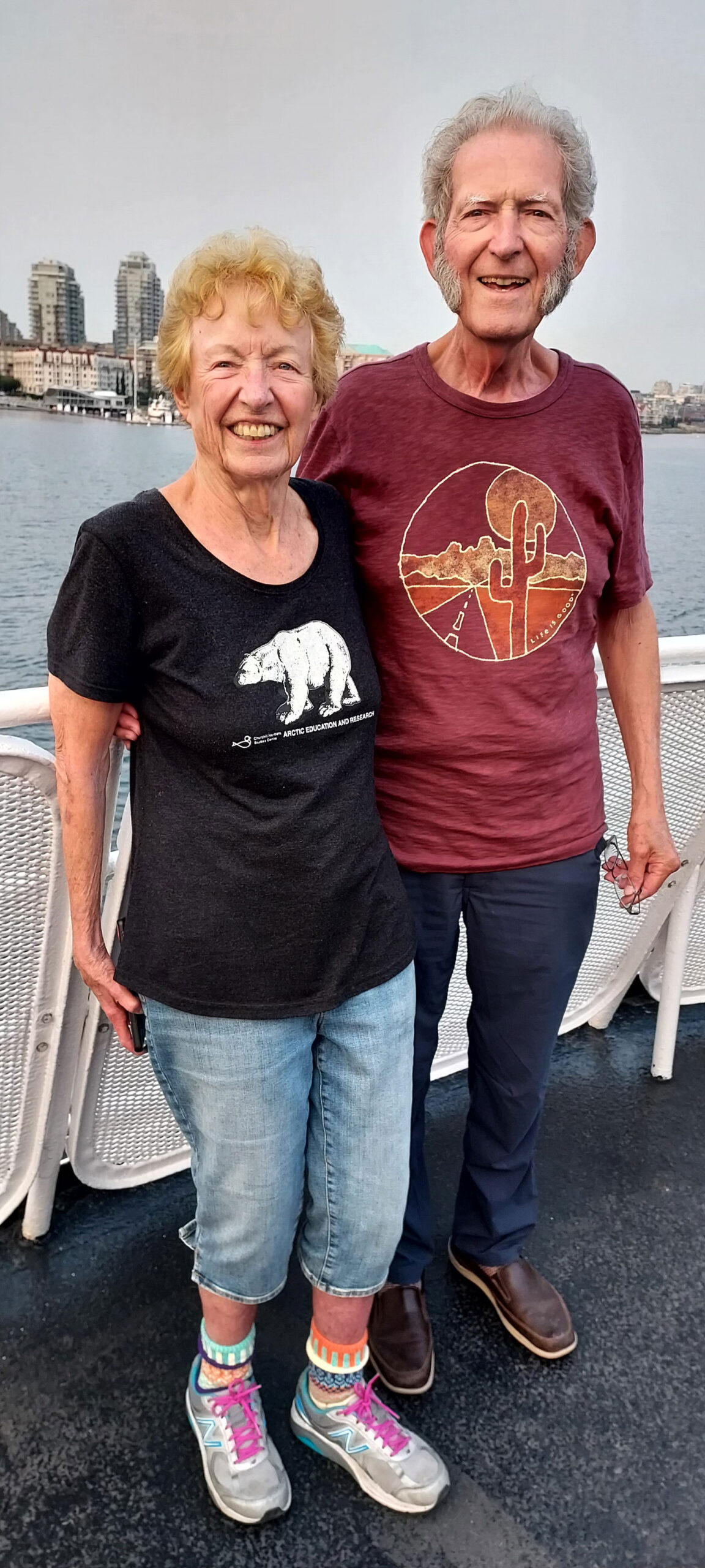 Sue Fairbanks and Jim Huff stand next to the water. A city skyline is in the background.