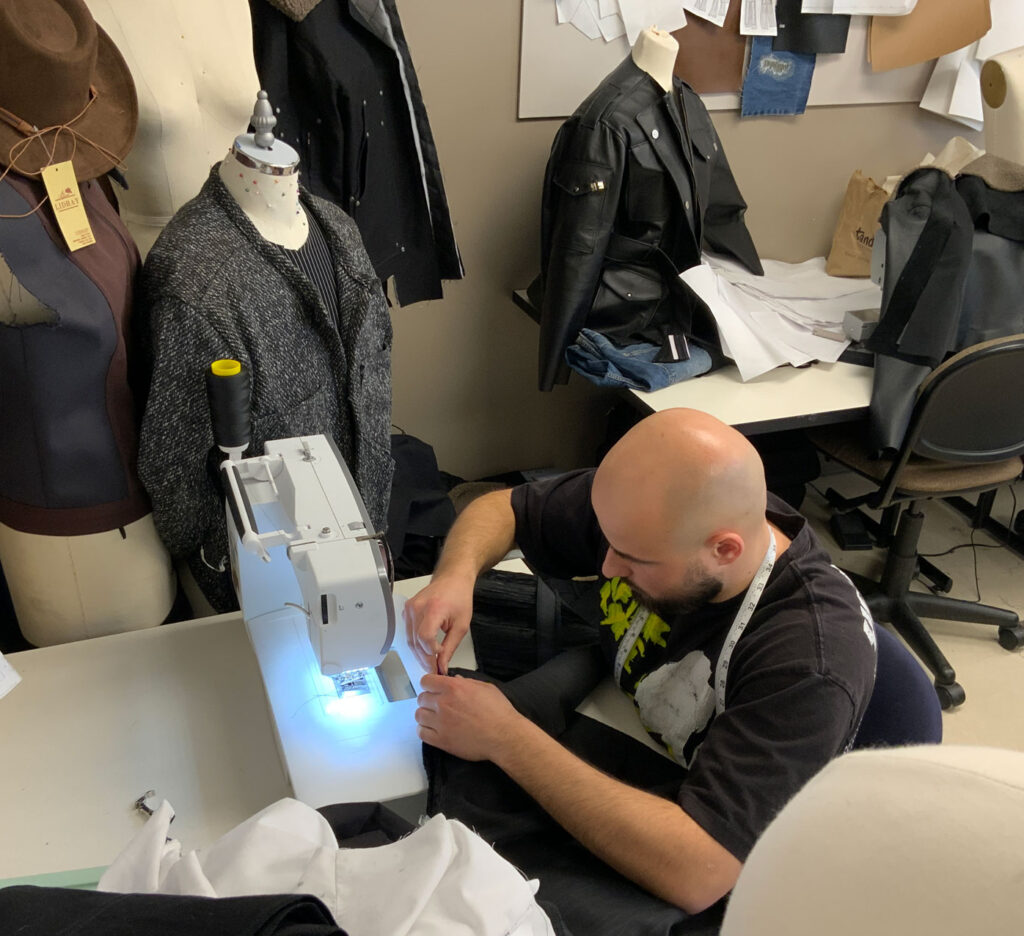 Kamil Hall works at a sewing machine. He is surrounded by mannequins with clothing on them.