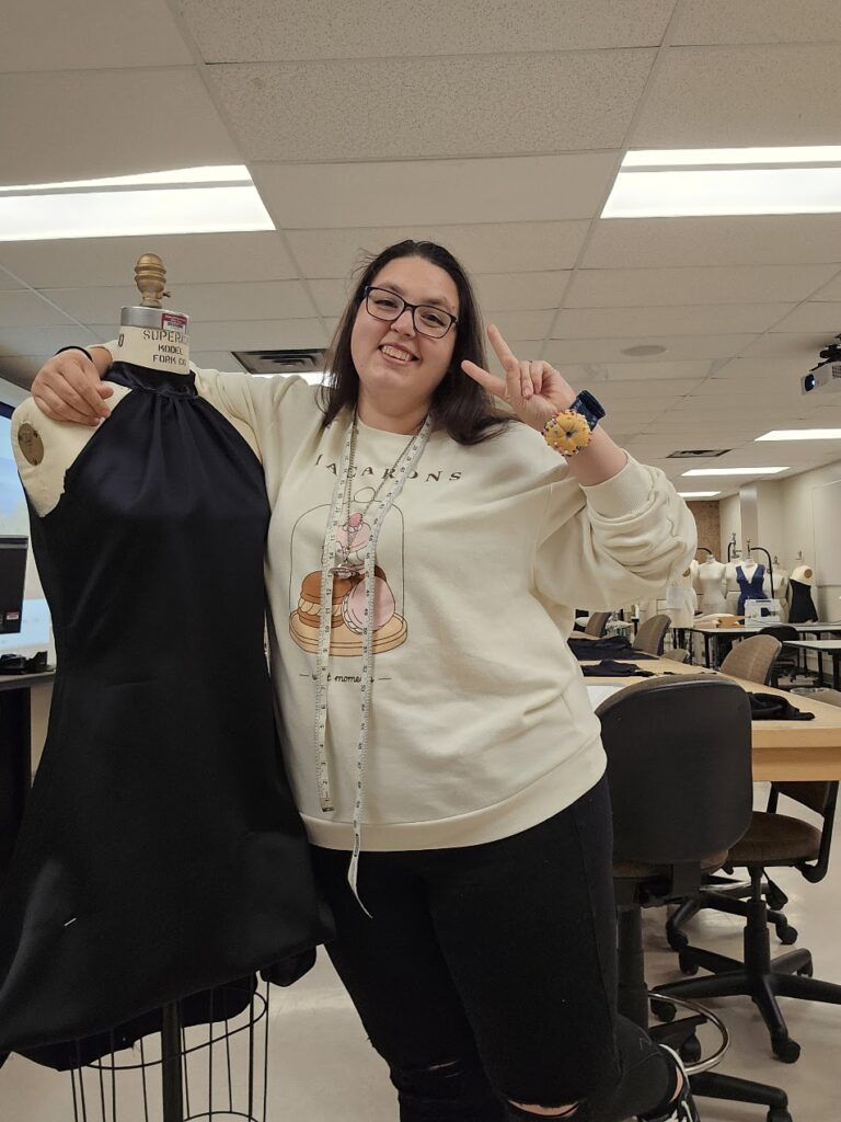 Kiah Conway poses with her arm around a mannequin with a dress on it while making the peace sign. Around Kiah's neck is a measuring tape.